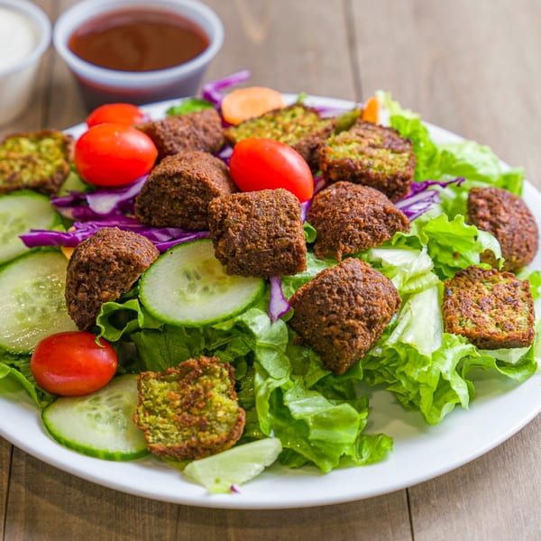 A vibrant halal falafel salad with fresh lettuce, cucumbers, tomatoes, and two sauces on a wooden table.
