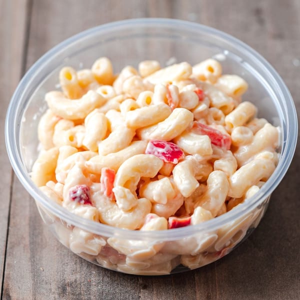 Halal macaroni salad with red peppers in a clear plastic container on a wooden surface.